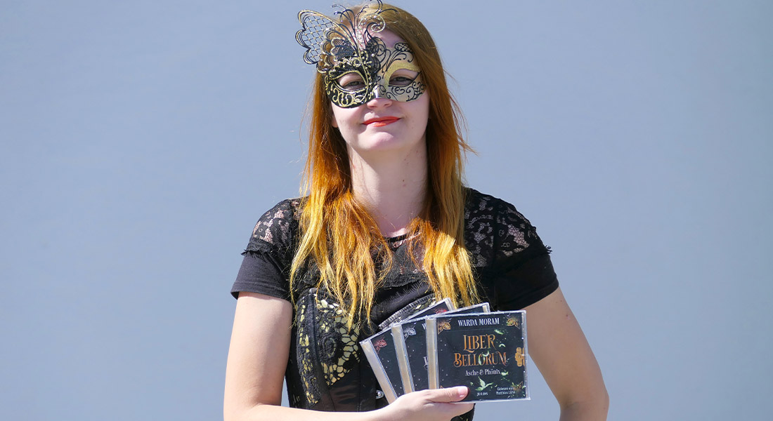 junge Frau mit Halbmaske hält 3 Hörbuch-CDs vor blauem Himmel